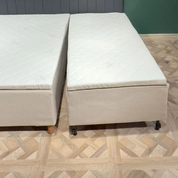 Du surinkti lovos moduliai stovi vienas šalia kito ant šviesių medinių grindų, tarp jų paliktas siauras tarpas. Kiekvienas modulis turi atskirą smėlio spalvos užvalkalą, dygsniuotą čiužinį ir kojeles.